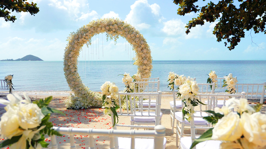 Beachfront destination wedding ceremony with circular white floral arch, chiavari chairs, and turquoise ocean backdrop